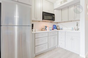 A kitchen with white cabinets, a stainless steel refrigerator, a black microwave, and various kitchen items on the counter. - Wade 'N Sea Condo with Pools & Hot Tubs (Galveston)