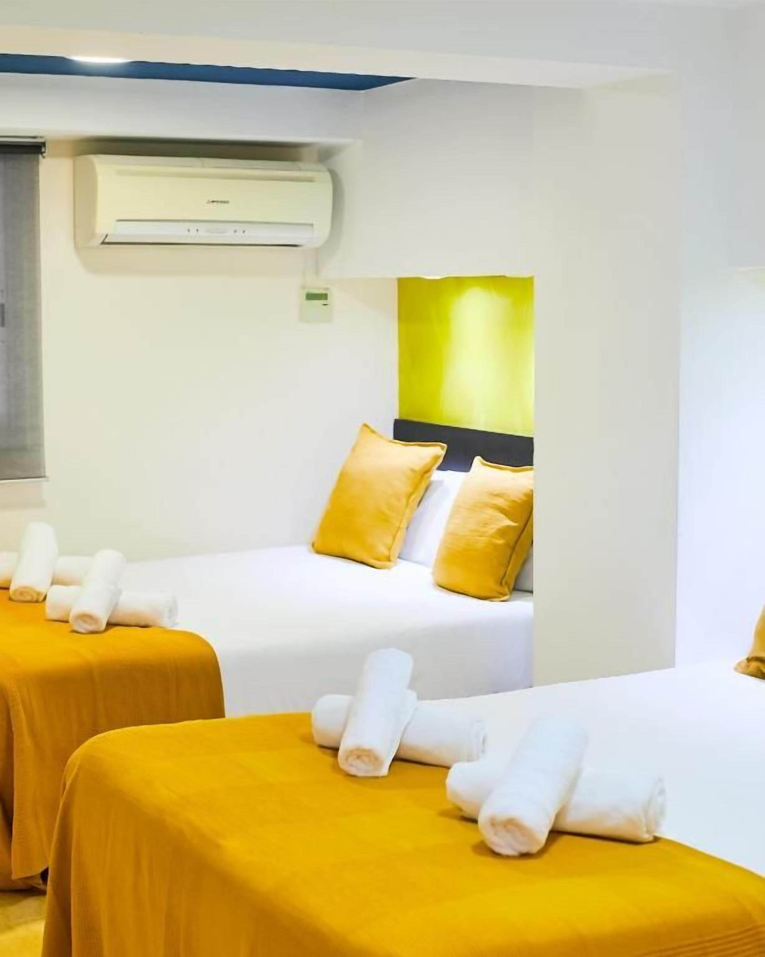 Habitación de hotel con dos camas, cada una con cubrecama amarillo y almohadas blancas, y un aire acondicionado de pared.