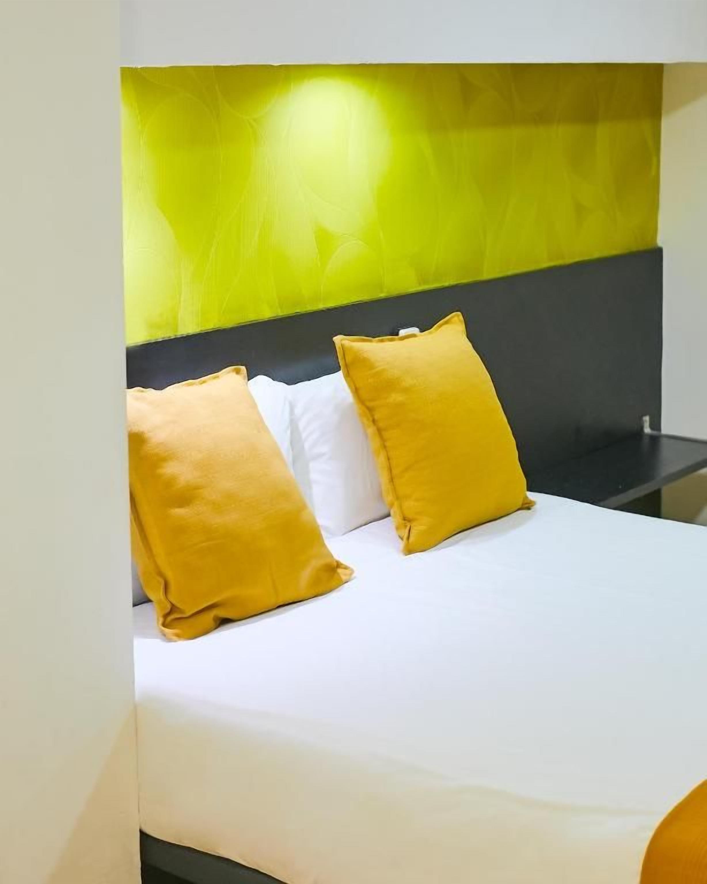 A bed with white bedding and two yellow pillows, a black headboard, and a green accent wall.