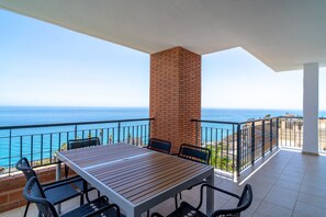 Ein Balkon mit Holztisch und -stühlen, einer Ziegelstütze und Blick auf das Meer.