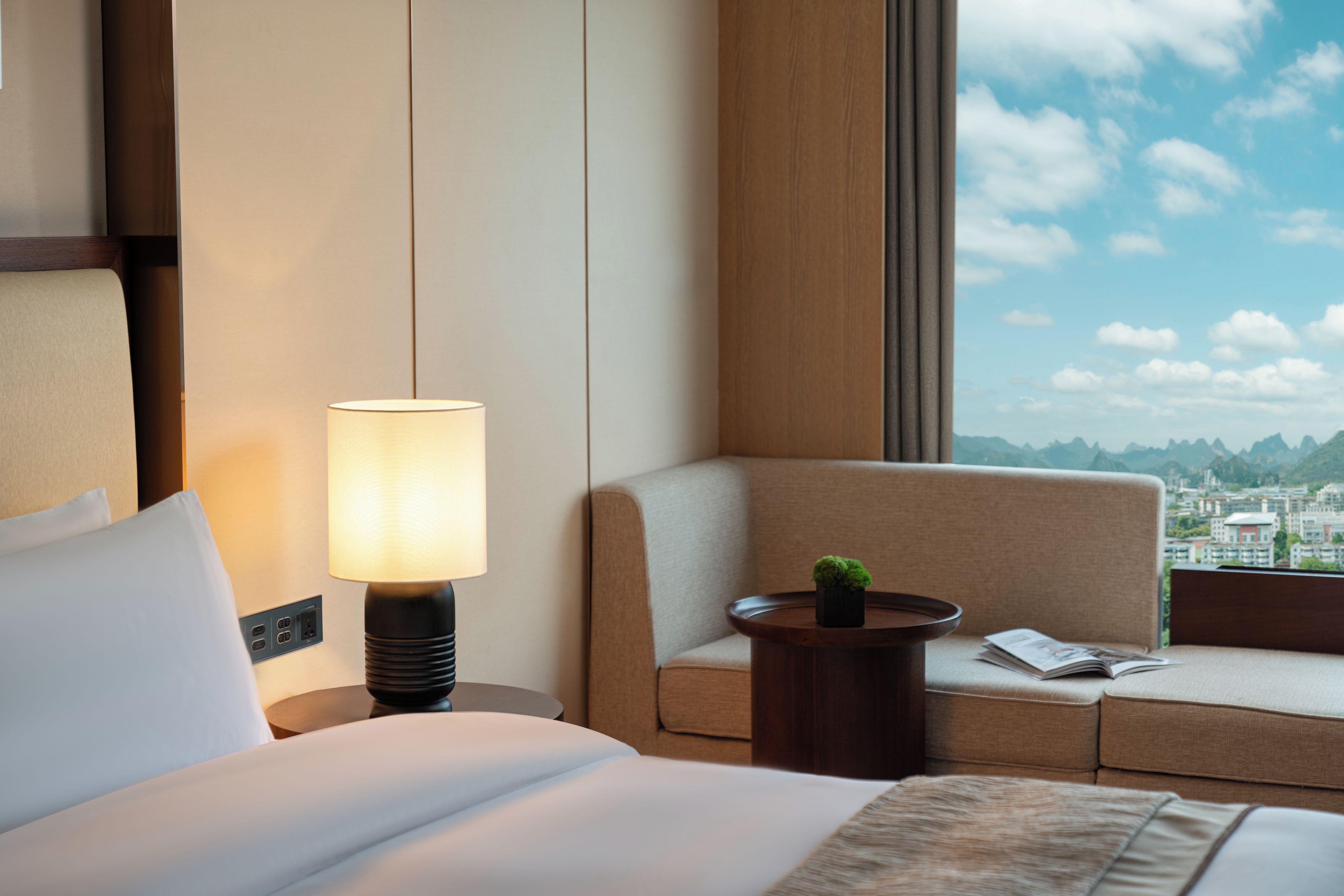A hotel room with a bed, a bedside lamp, a small table with a plant, a sofa, and a window with a city view.