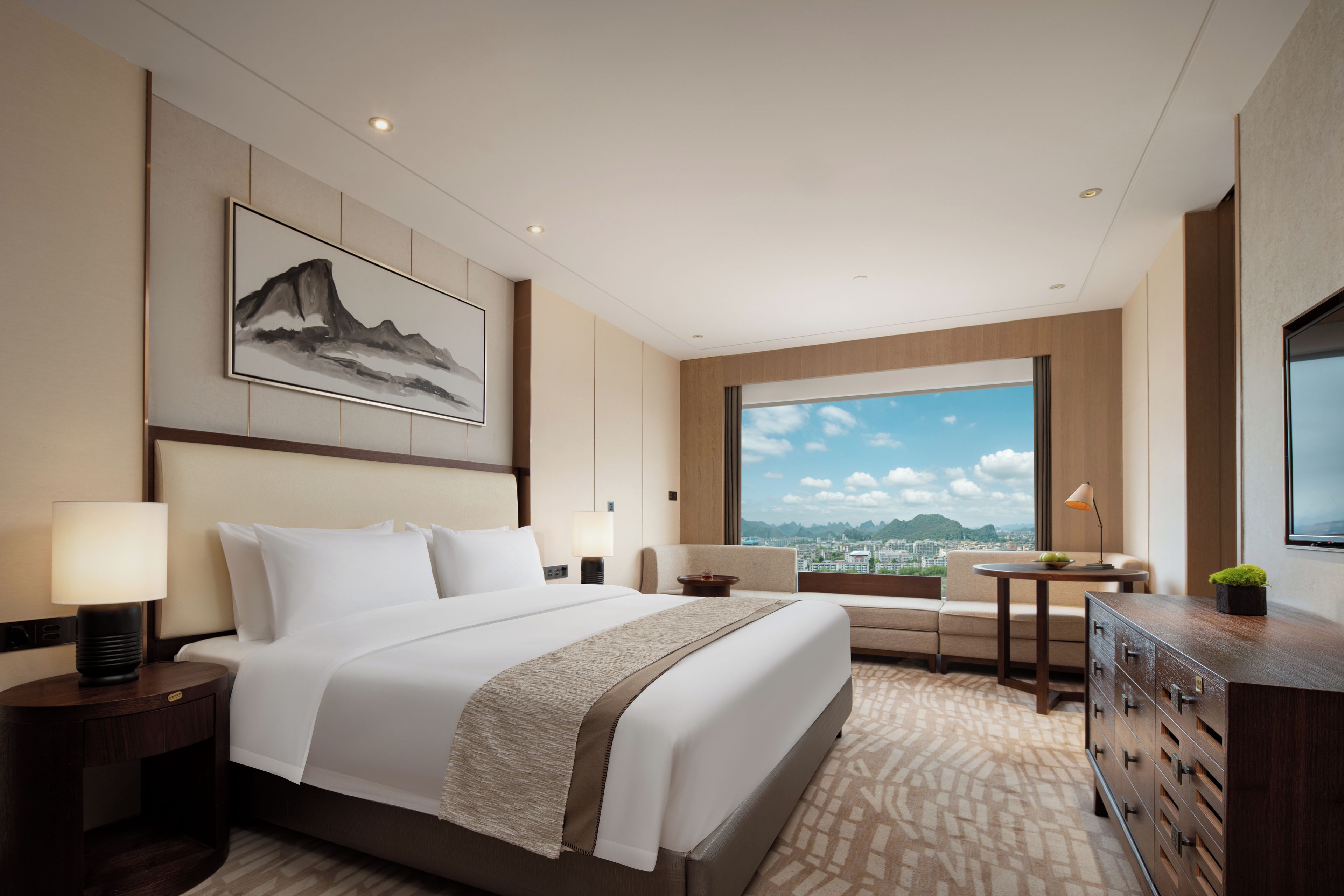 A hotel room with a large bed, a desk, a chair, a TV, and a window with a city view.