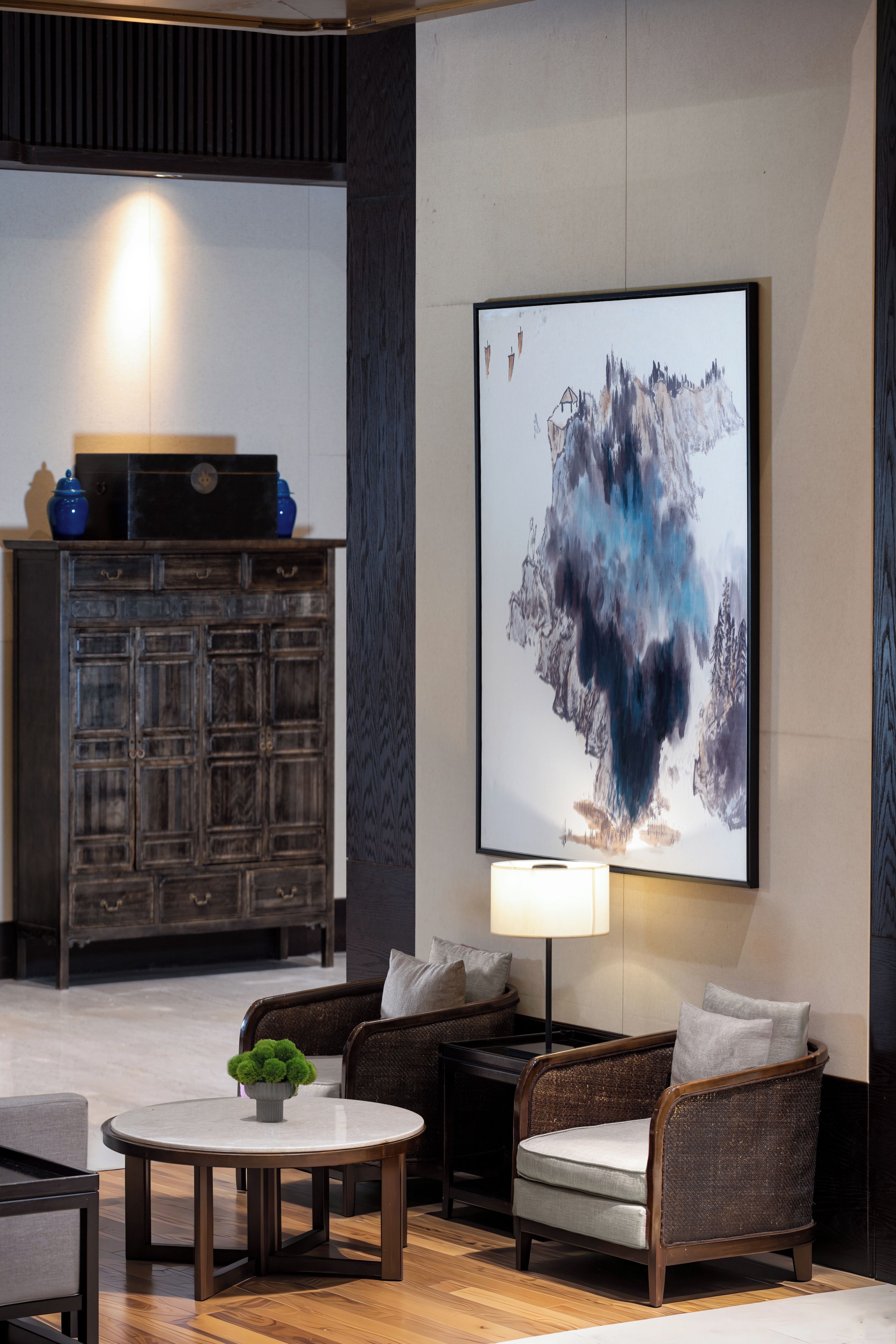 Un área de descanso moderna con un cuadro abstracto grande, un mueble de madera oscura, mobiliario de mimbre y una mesa redonda con un florero.