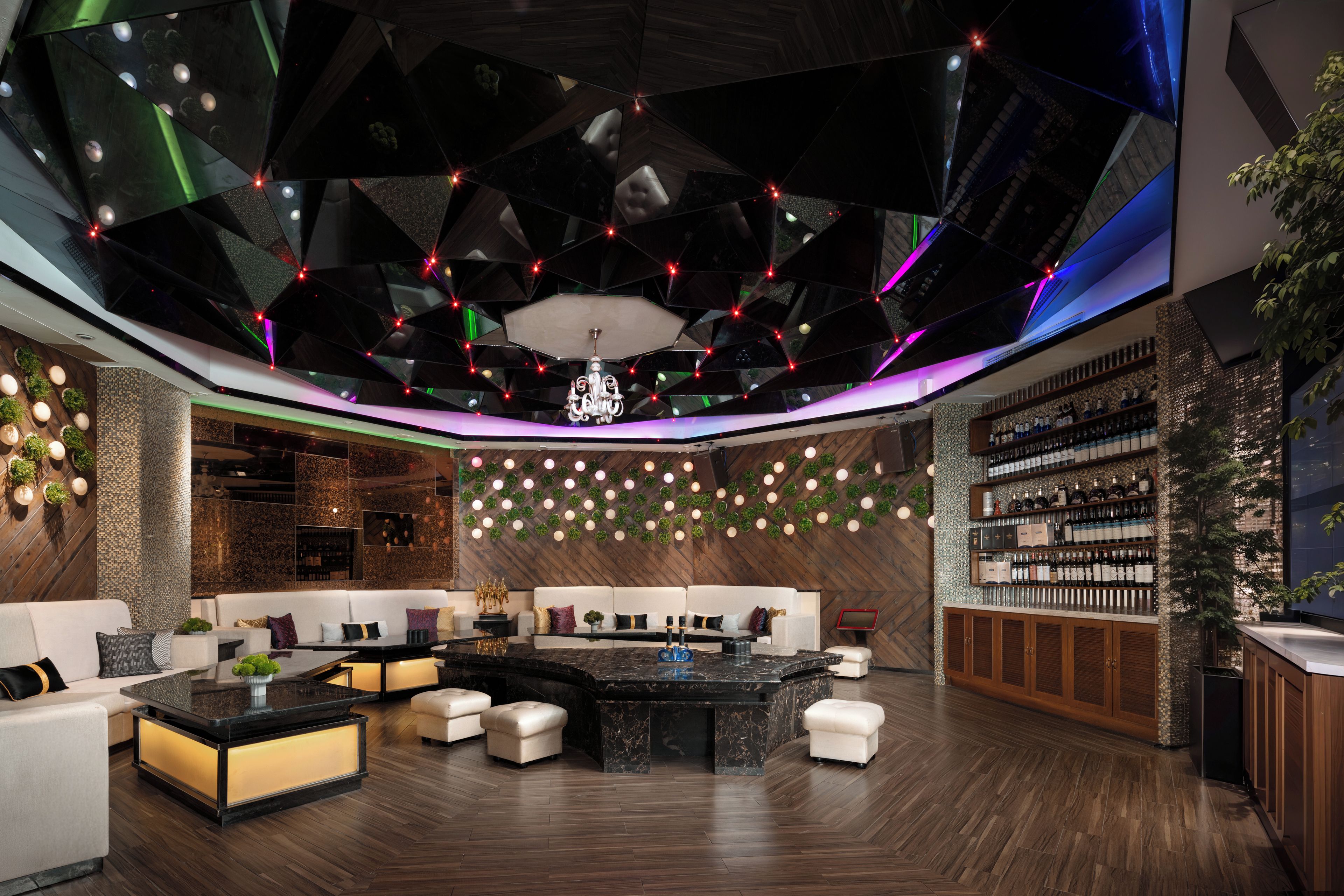 Un espace lounge moderne avec un aménagement central autour d’un coin salon, un bar et un mur entier recouvert d’étagères remplies de bouteilles.