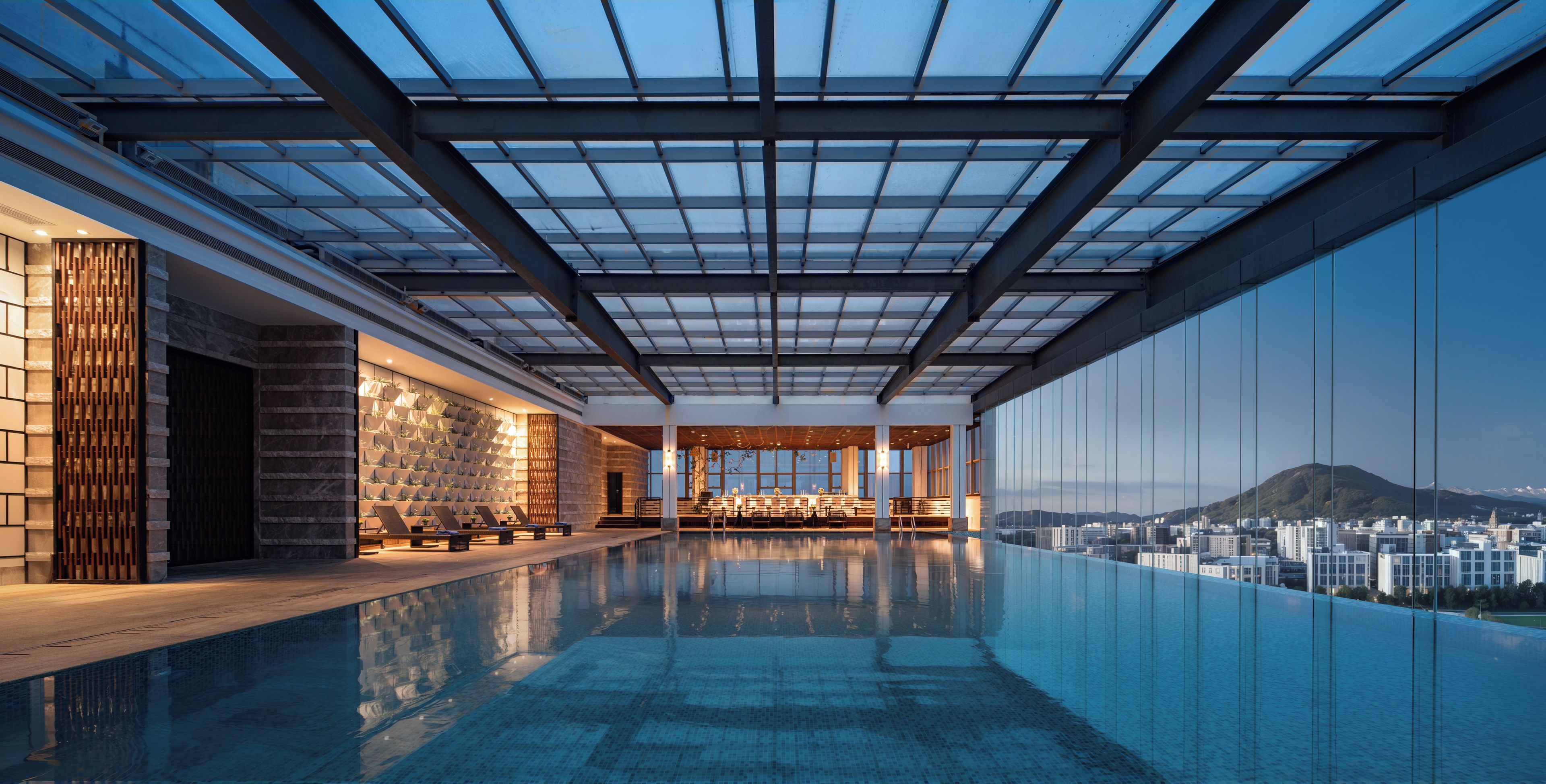 Une piscine à débordement moderne avec un toit en verre, un espace salon agrémenté de plantes et une vue sur la ville.