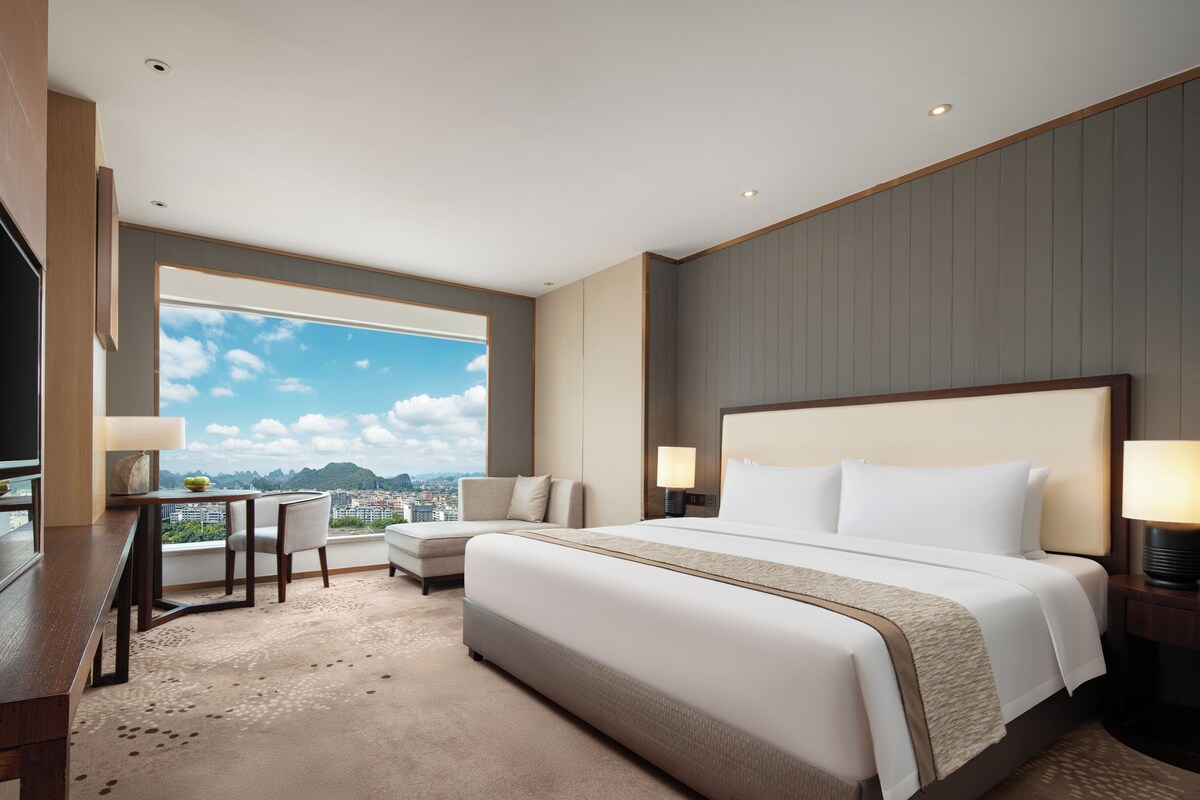 A hotel room with a large bed, a desk, a chair, and a view of a cityscape.