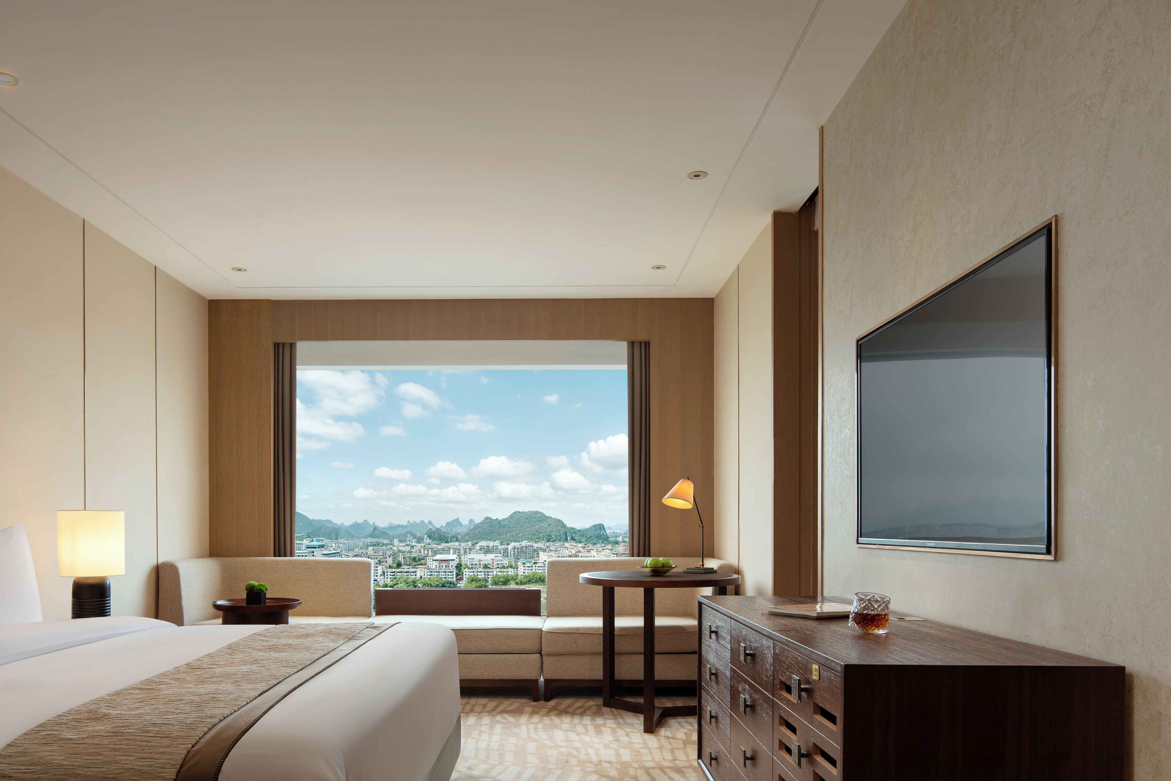 Ein Hotelzimmer mit einem großen Bett, einem Schreibtisch, einem Sessel, einem Fernseher und Blick auf eine Stadtlandschaft.