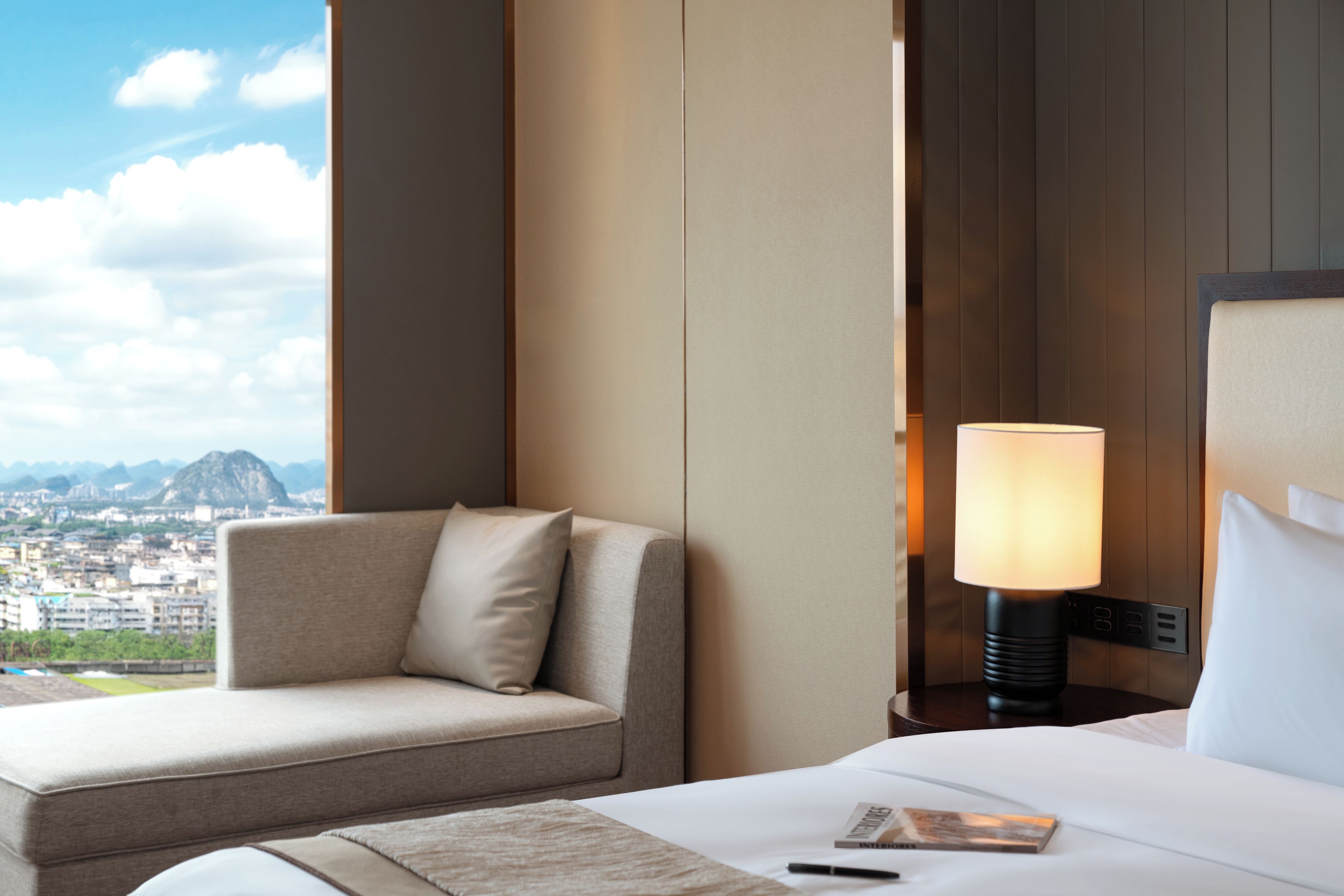 Ein Hotelzimmer mit Bett, Nachttisch mit Lampe, Sofa und Blick auf eine Stadtlandschaft.