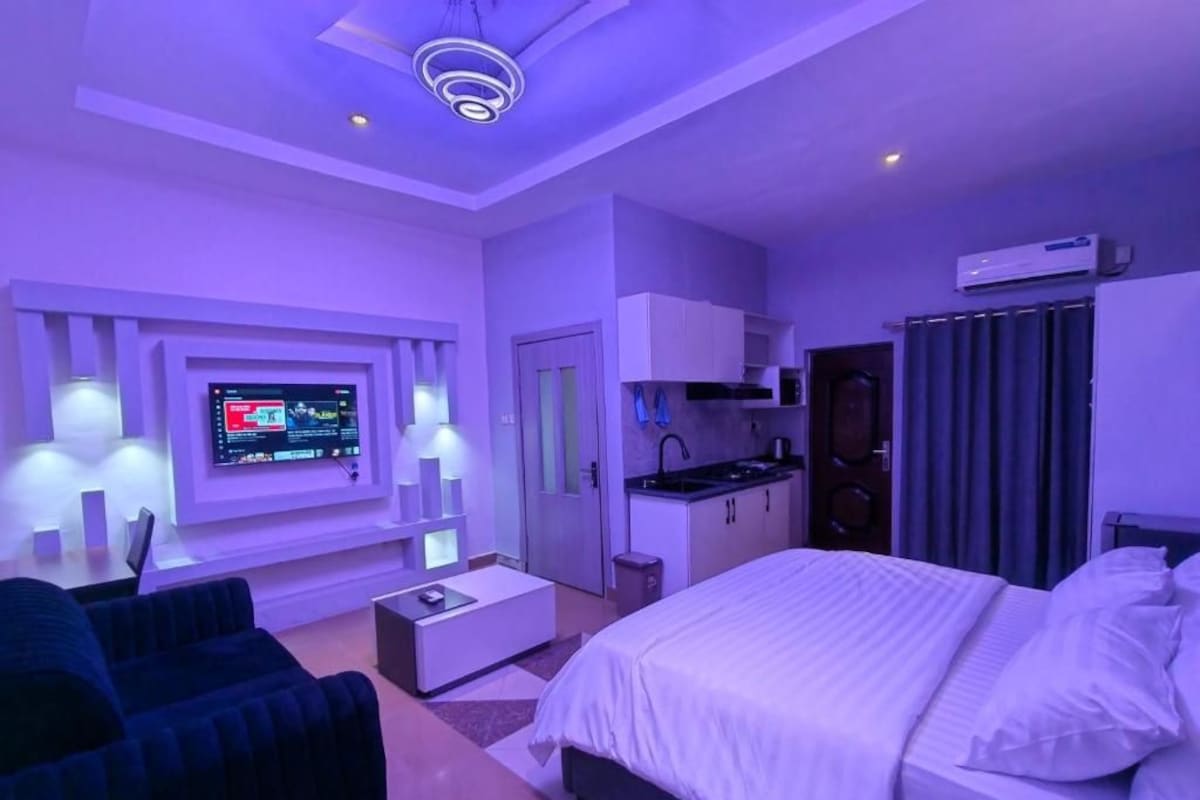 A modern hotel room with a bed, a TV mounted on the wall, a kitchenette, and a seating area with a sofa.