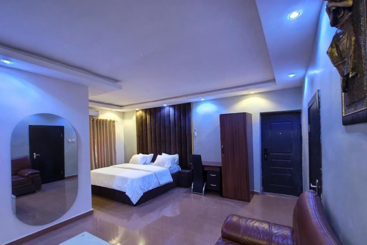 A modern hotel room with a large bed, a seating area, a desk, and a mirror.