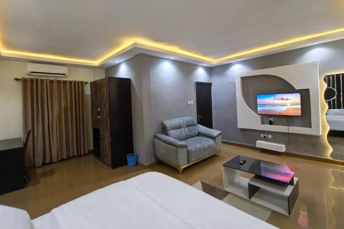 A modern hotel room with a bed, a sofa, a TV mounted on the wall, and a ceiling with recessed lighting.