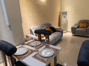 A dining area with a table set for two, a grey sofa, and a modern air conditioning unit in the background. - Milestone Boutique Apartments (ikeja)