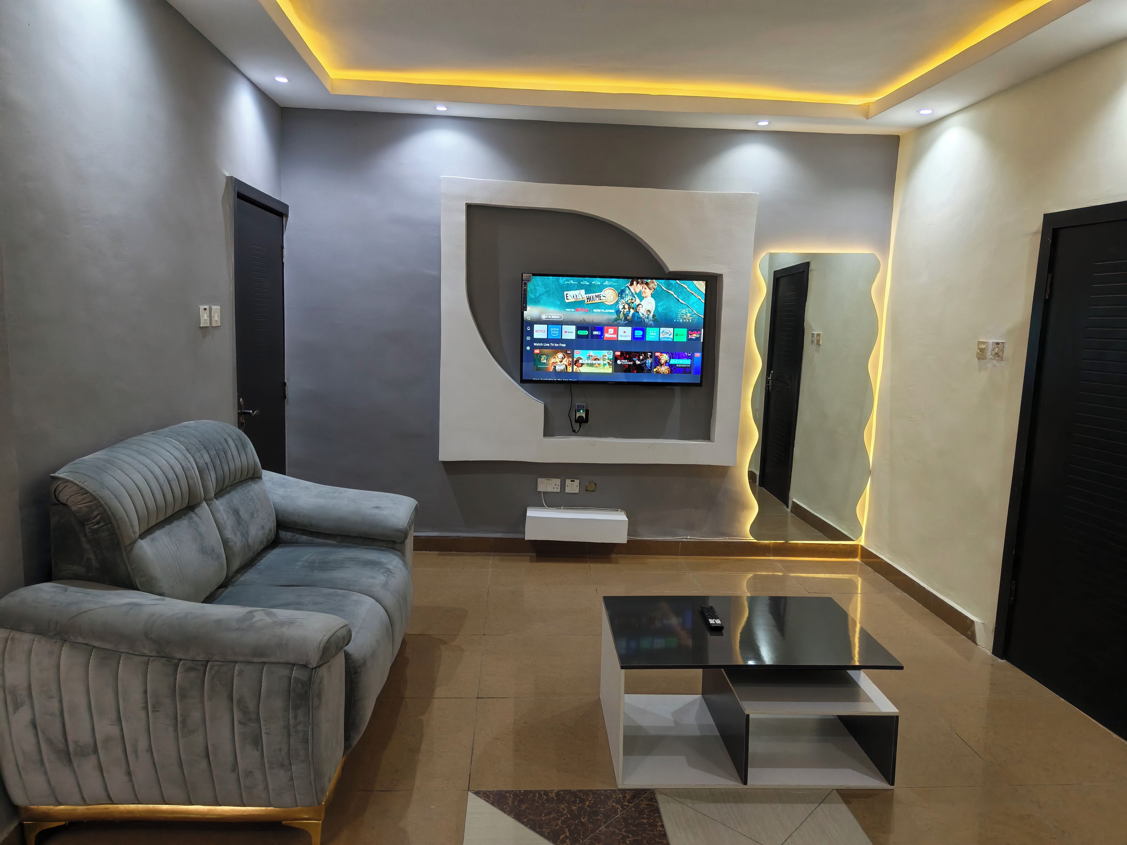 Un salon moderne avec un canapé gris, un téléviseur à écran plat fixé au mur et une table basse en verre.