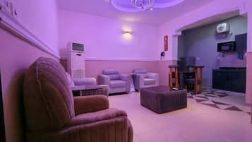 A living room with a sofa, armchair, and ottoman, a ceiling light fixture, and a kitchen area with a microwave.