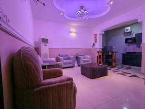 Sala de estar con sofá, sillón y reposapiés, una lámpara de techo y una zona de cocina con microondas.