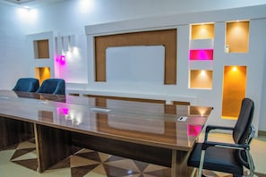 Sala de conferências com uma mesa comprida, cadeiras, quadro branco e iluminação fixa na parede.