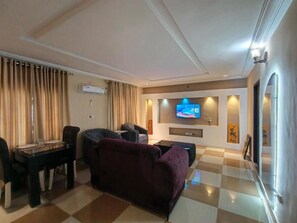Habitación de hotel con piso a cuadros, un sofá, una mesa de comedor con sillas, un televisor de pantalla plana montado en la pared y un equipo de aire acondicionado.