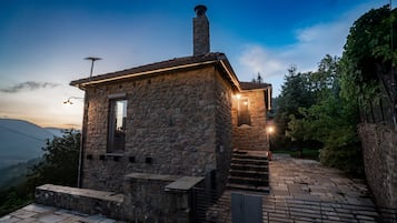 Edificio de piedra con chimenea, una ventana pequeña y una escalera que lleva a una puerta.