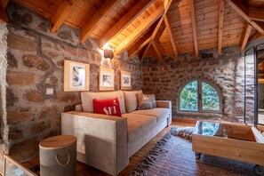 A cozy room with a stone wall, wooden ceiling beams, a bed with a red pillow, a small table, and a window with a view of trees.