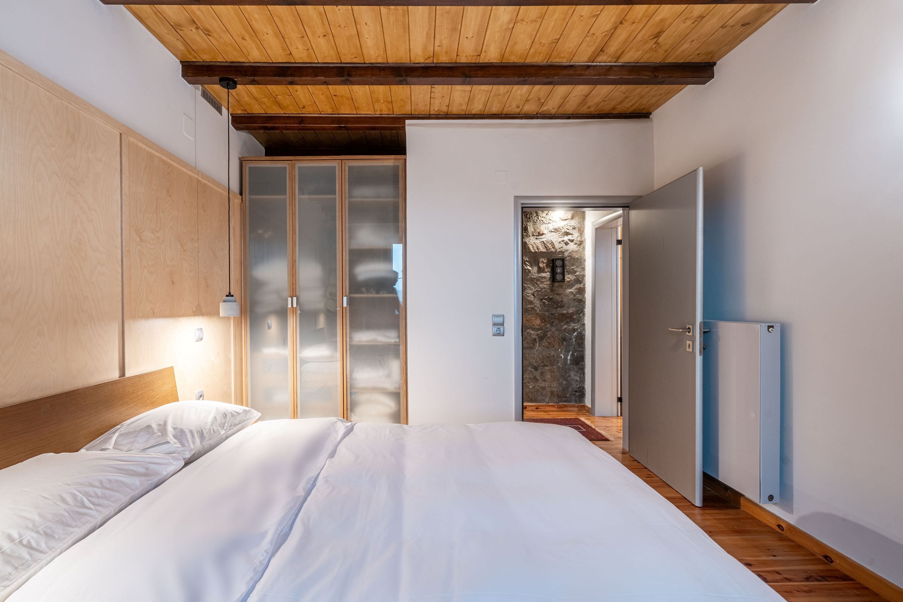 Un dormitorio con techo de madera, cama con ropa blanca, armario y puerta que da a un balcón.