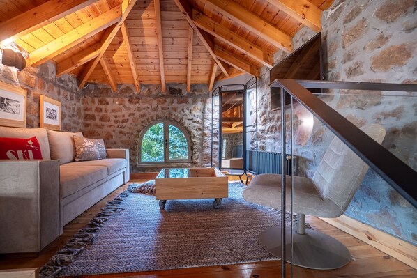 A cozy attic room with a stone wall, wooden beams, a wooden sofa, a glass partition, and a view through a window.