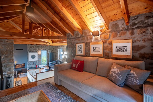 A cozy living room with a stone wall, wooden beams, a sofa with cushions, and framed pictures.