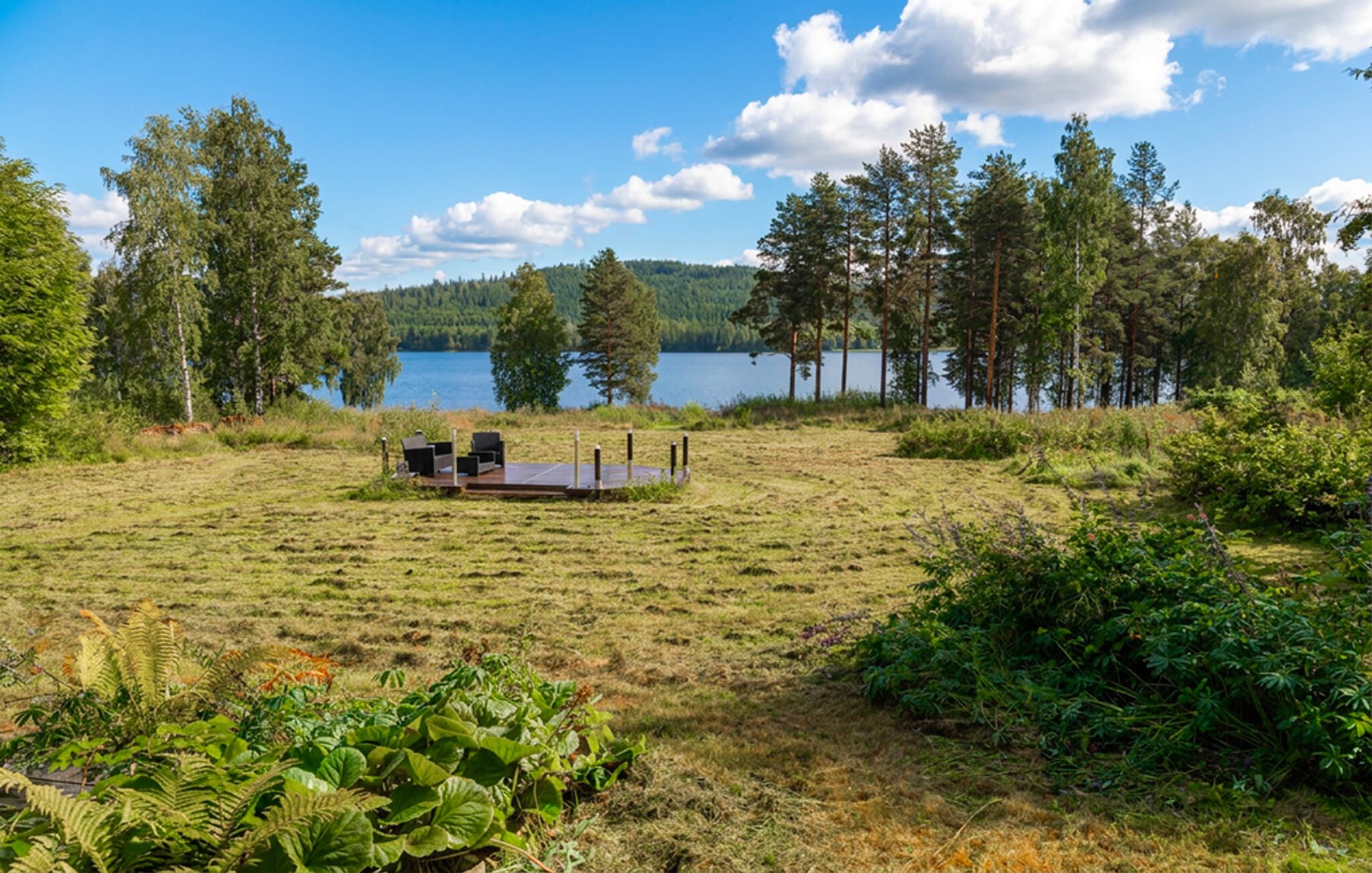 Ett utomhusområde med sittplatser vid sjön, med ett trädäck och omgivet av träd och växtlighet.