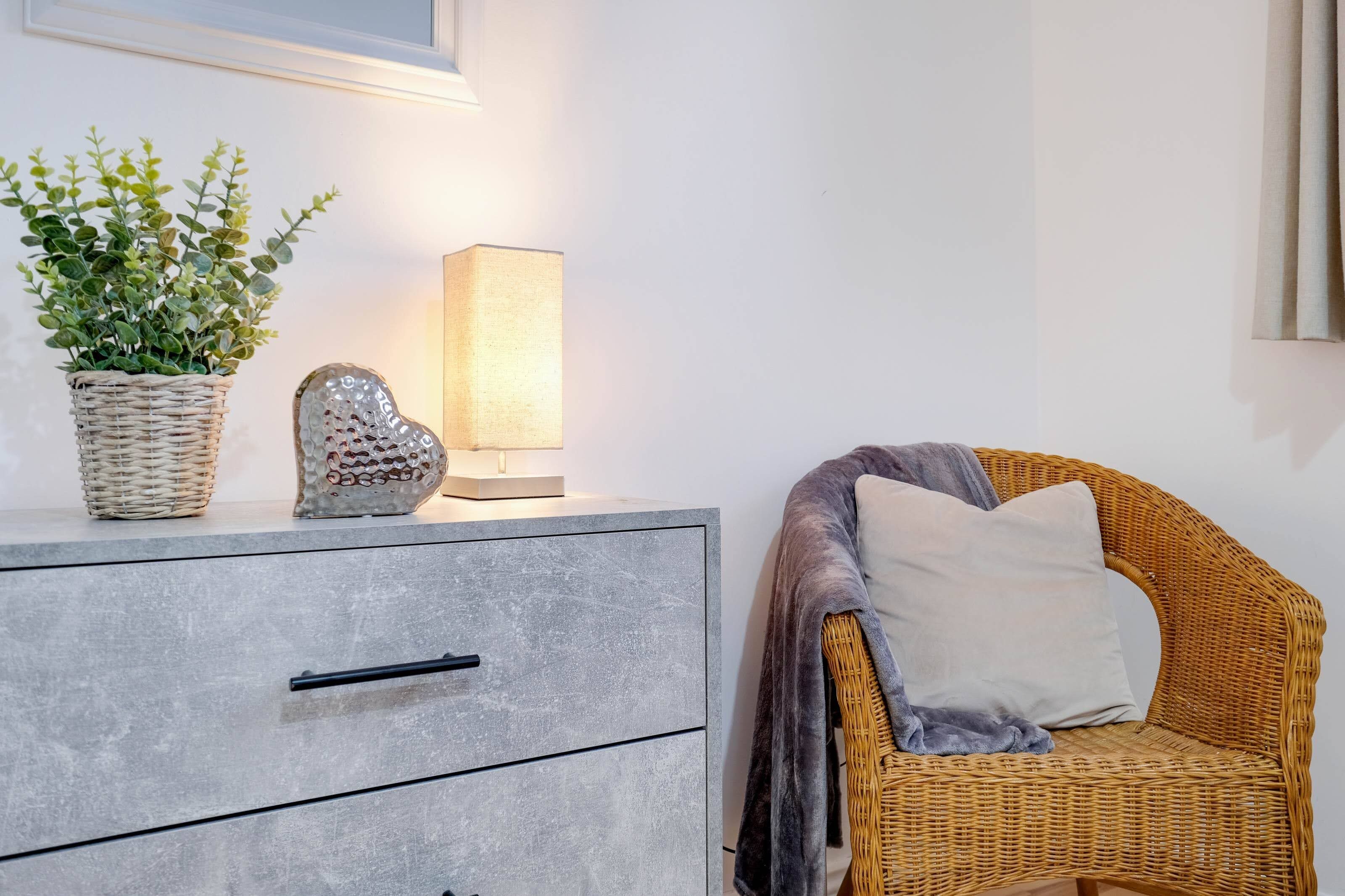 A room with a grey drawer unit, a wicker chair with a cushion, a potted plant, and a wall-mounted lamp.