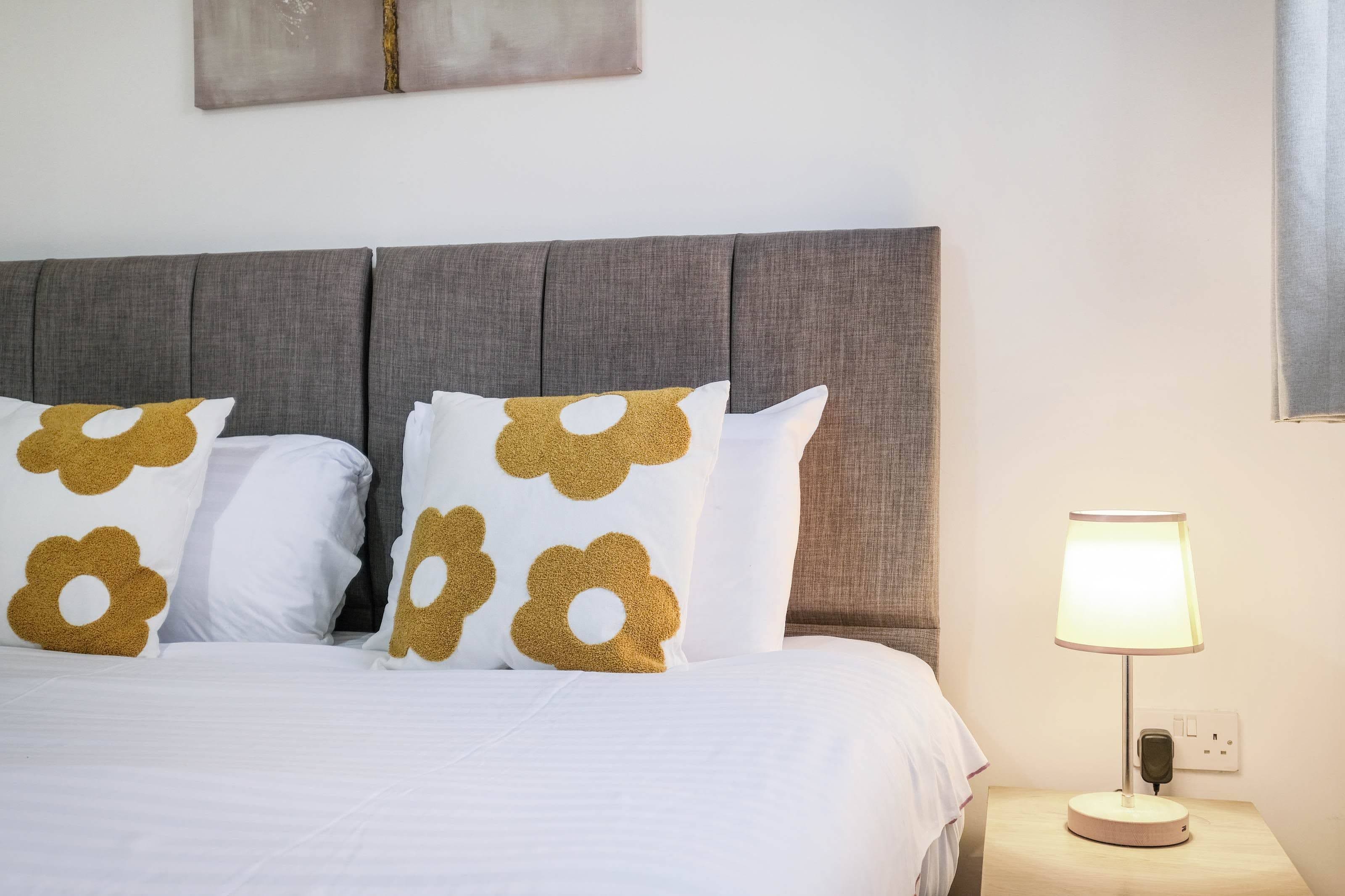 A neatly made bed with a grey upholstered headboard, white bedding, and decorative pillows with a gold pattern.