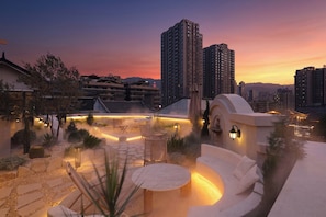Un jardín en la azotea con zonas de estar, fuente de agua y vistas a edificios altos circundantes al atardecer.