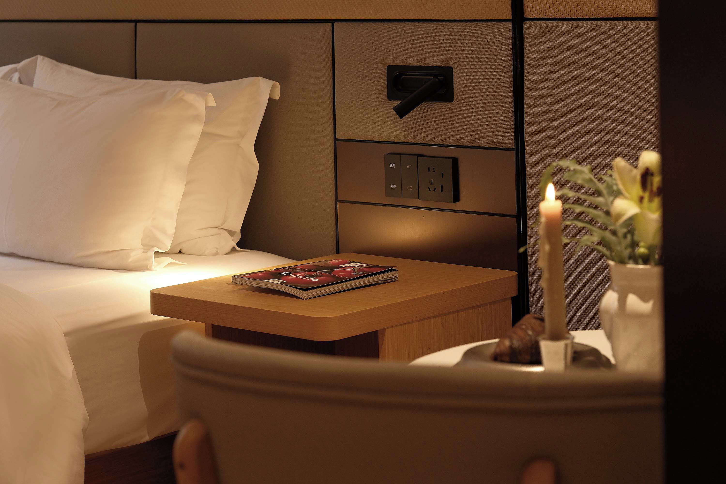 A hotel room with a neatly made bed, a bedside table with a magazine, a wall-mounted power outlet, and a lit candle.