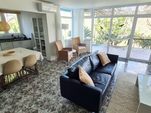 Sala de estar moderna com sofá de couro preto, mesa de jantar e cadeiras. Há uma cozinha com armários e janela com vista para um jardim.