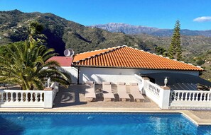 Área de piscina com um edifício branco, telhado de telhas vermelhas e montanhas ao fundo.