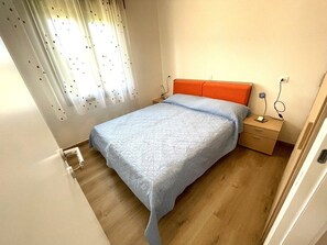 Une chambre à coucher avec un lit, une table de chevet, une prise de courant et une fenêtre avec des rideaux.