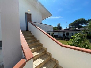 Eine Treppe führt zu einem Gebäudeeingang mit einer Tür, umgeben von einer niedrigen Mauer und einem kleinen Garten.