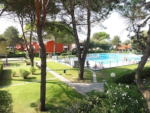 A swimming pool surrounded by trees and a grassy area with a red building in the background. - Accommodation in residence with pool and garden (Bibione)
