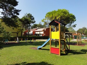 A playground with a slide and climbing structures, surrounded by grass and trees, with a building in the background. - First-floor accommodation in residence with pool (Bibione)
