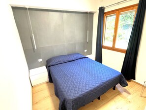A single bed with a quilted blue bedspread in a room with a wooden floor, a window with curtains, and a wall-mounted headboard. - Holiday home with garden in Bibione Pineda (Bibione Pineda)