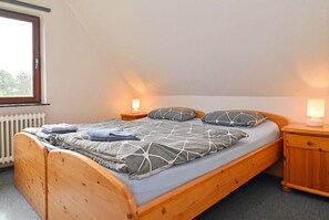 Ein Einzelbett mit Holzrahmen und einer grau gemusterten Bettdecke, flankiert von zwei Nachttischen mit Lampen, in einem Zimmer mit schrägen Wänden und einem Fenster.