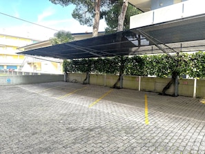 A covered parking area with a paved surface, a wall with a tree, and a building in the background. - Central flat 250m from beach (Bibione)