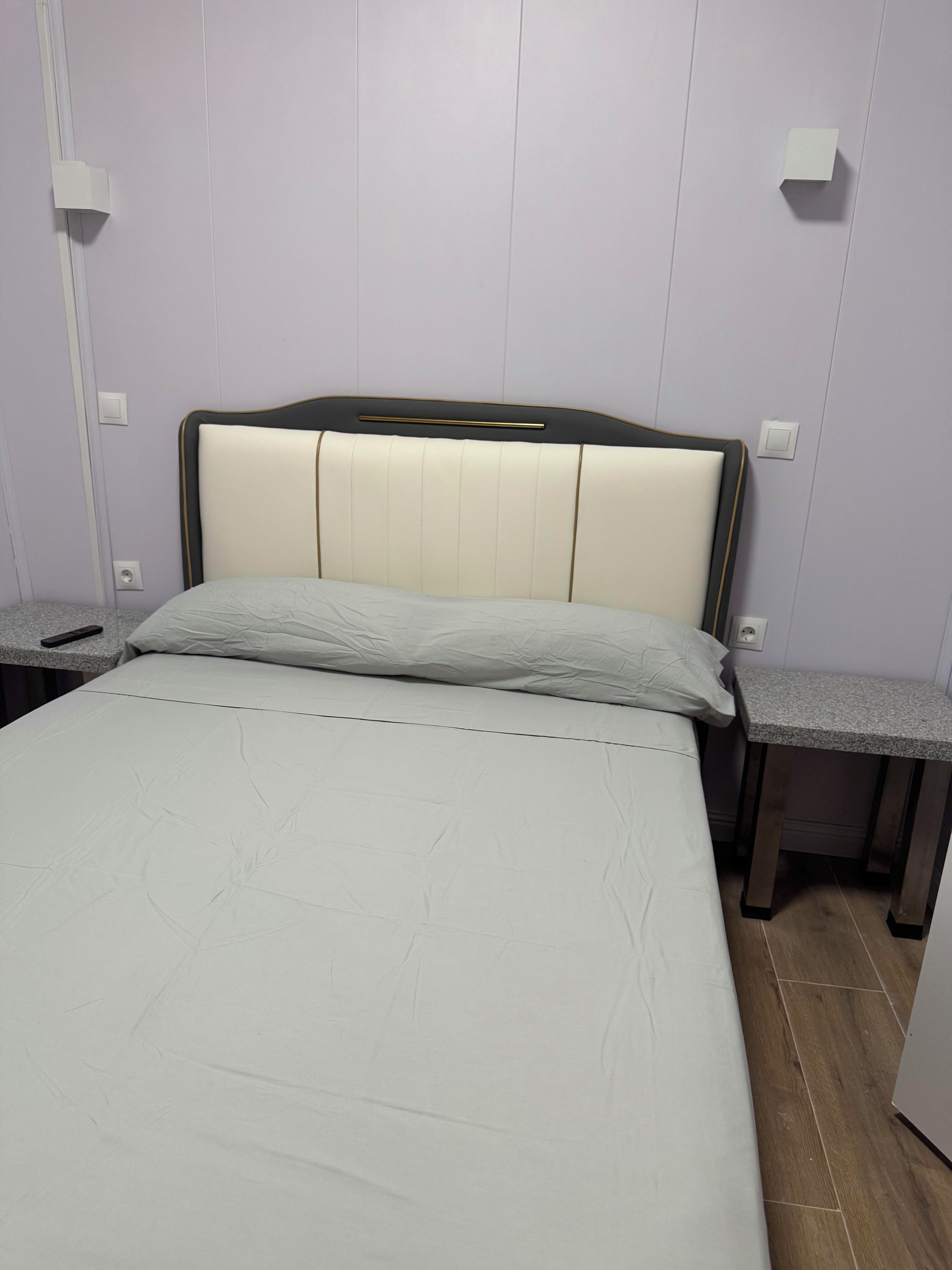 Een bed met hoofdeinde, twee nachtkastjes en een lichtschakelaar aan de muur.