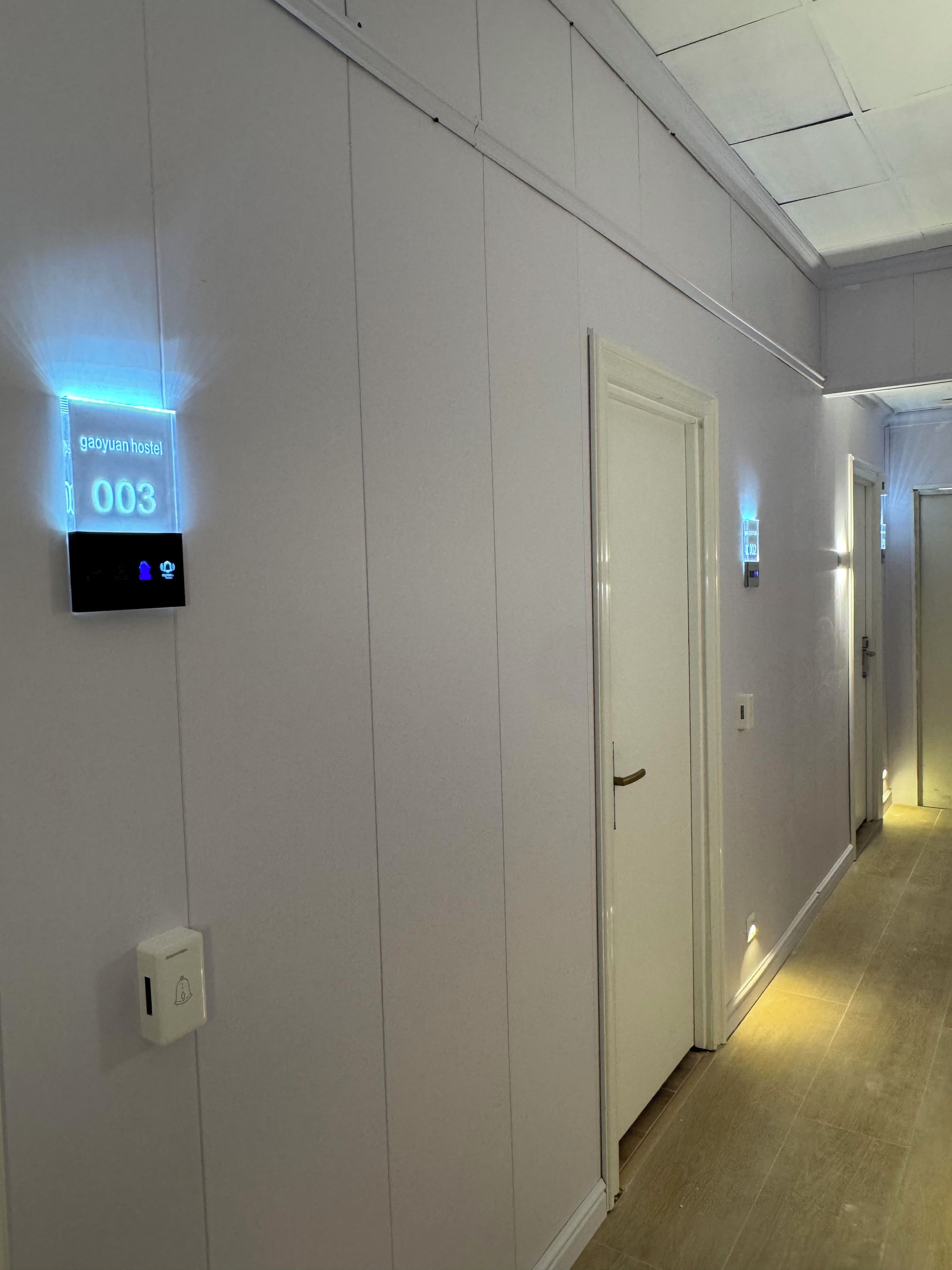 Een gang met een digitaal bord waarop kamer 003 staat, een deur en een lichtschakelaar.