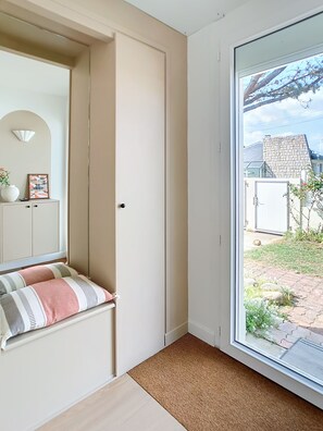 Ein Zimmer mit einer Schiebetür zu einem Garten, einer Bank mit Kissen, einem Schrank und einer Wandleuchte.