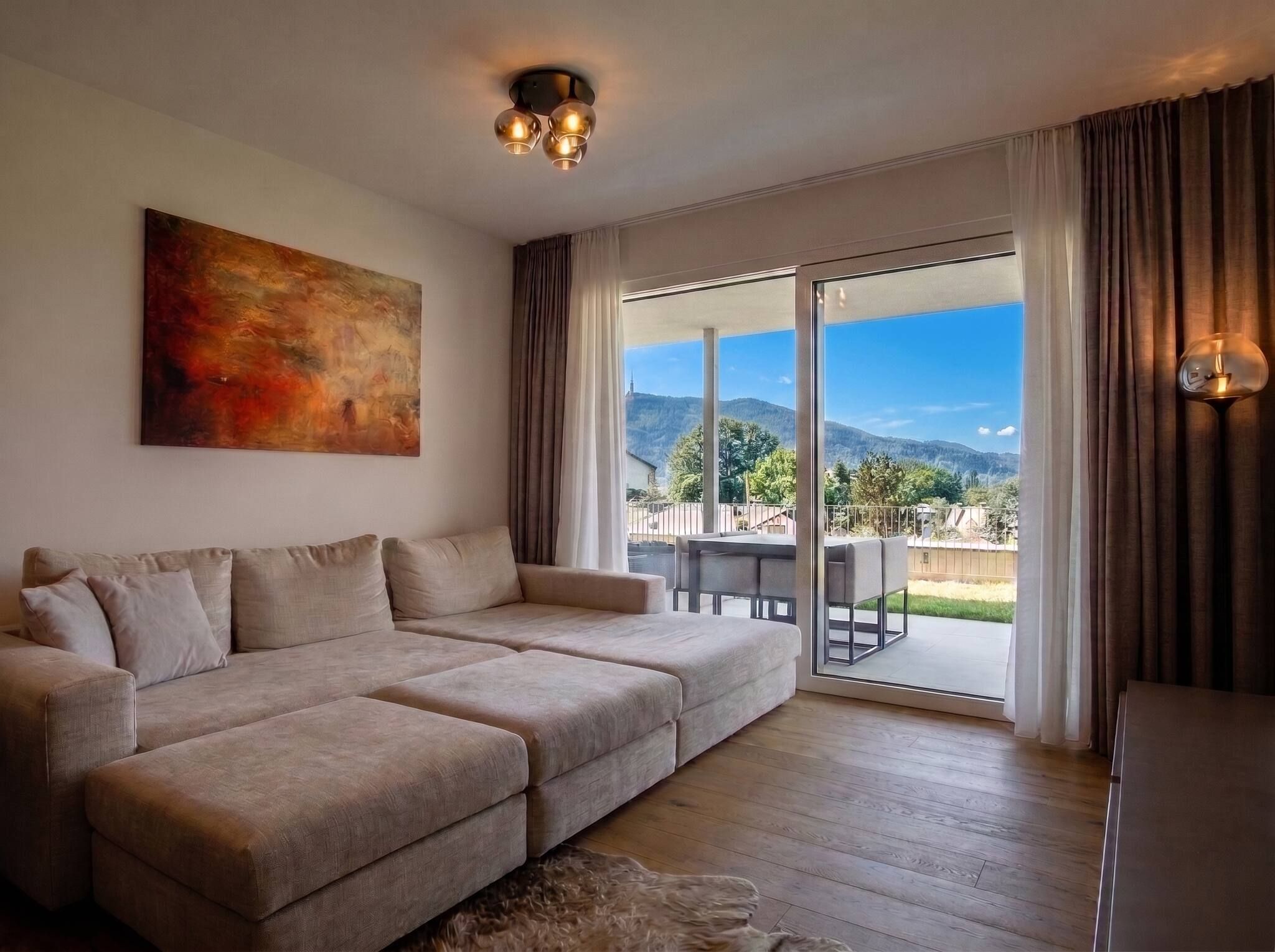 Ein Wohnzimmer mit einem beigefarbenen Ecksofa, einem Bild an der Wand und Blick auf eine Berglandschaft durch die Glasdoor.