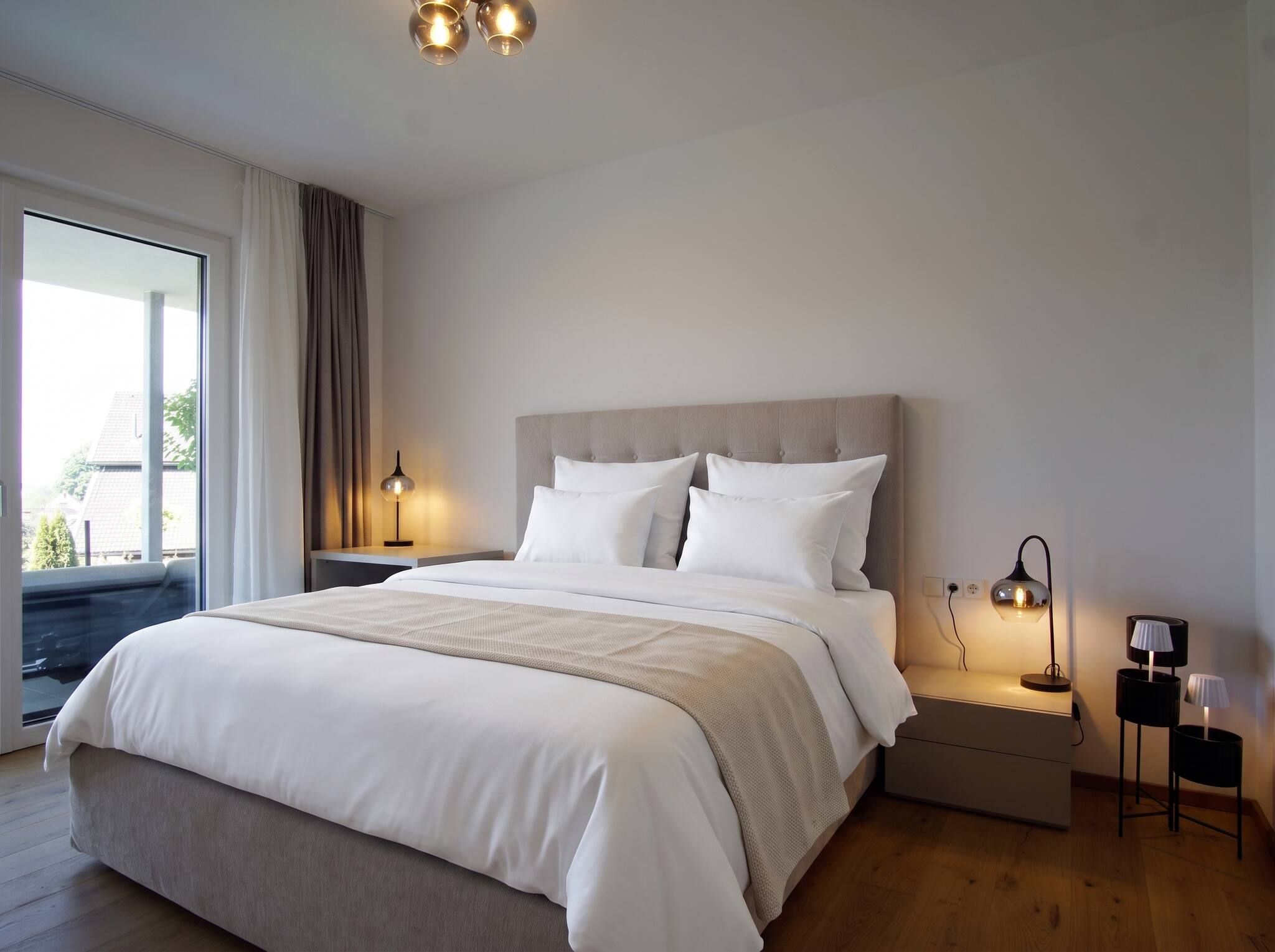 A modern bedroom with a large bed, two bedside lamps, and a view of the outdoors.