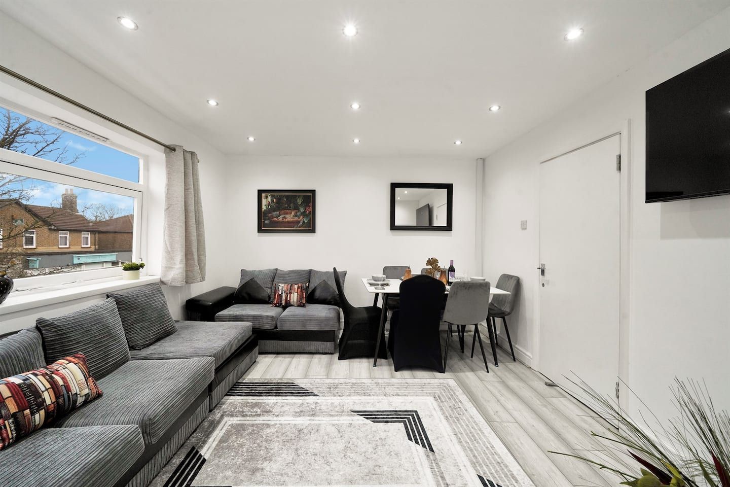 Ett modernt vardagsrum med en hörnsoffa, ett matbord med stolar, en platt-TV som är monterad på väggen och ett fönster med utsikt över ett bostadsområde.