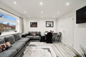 Una sala de estar moderna con un sofá modular, una mesa de comedor con sillas, un televisor de pantalla plana montado en la pared y un ventanal con vistas a una zona residencial.