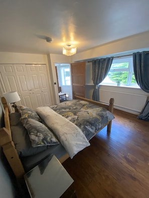 A bedroom with a bed, a bedside table, a lamp, a window with curtains, and a view of the outdoors. - Blissful Retreat - 4 Bedroomed House (Bridgend)