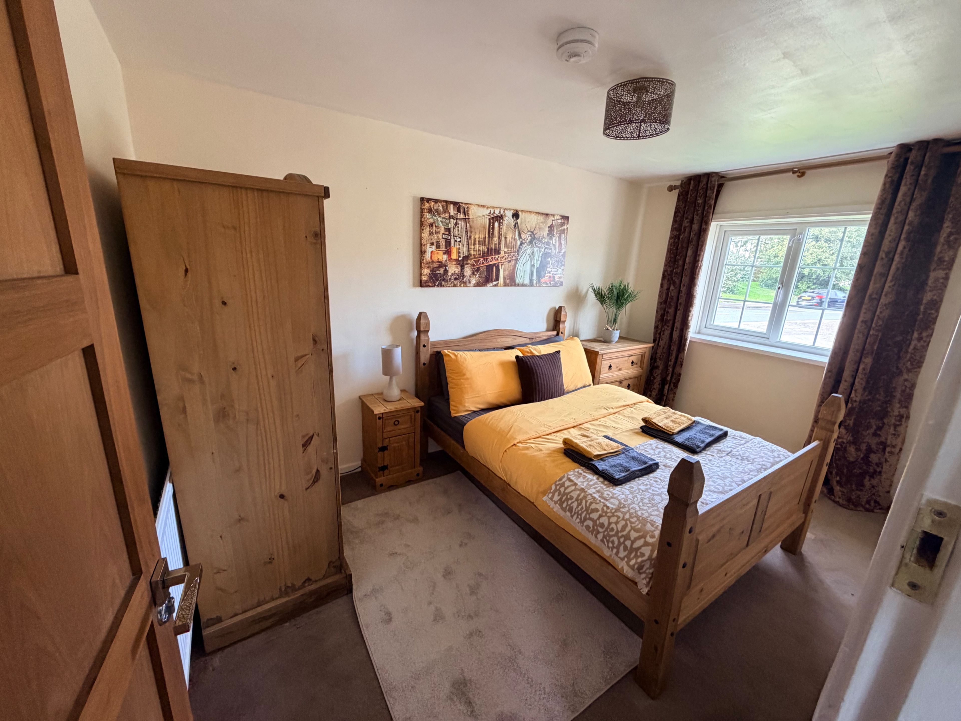 A bedroom with a wooden bed, a nightstand, a wardrobe, a window with curtains, and a ceiling light fixture.
