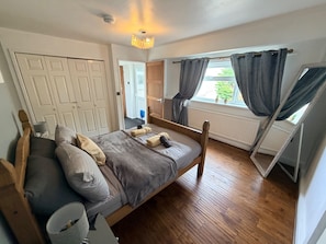 Un dormitorio con cama de madera, un radiador, una ventana con cortinas y una puerta que da a otra habitación.